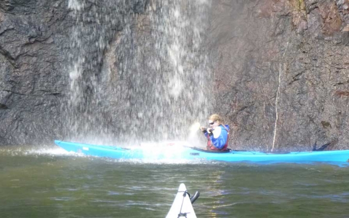 a person in a blue kayak paddles under a waterfall in front of a rock wall